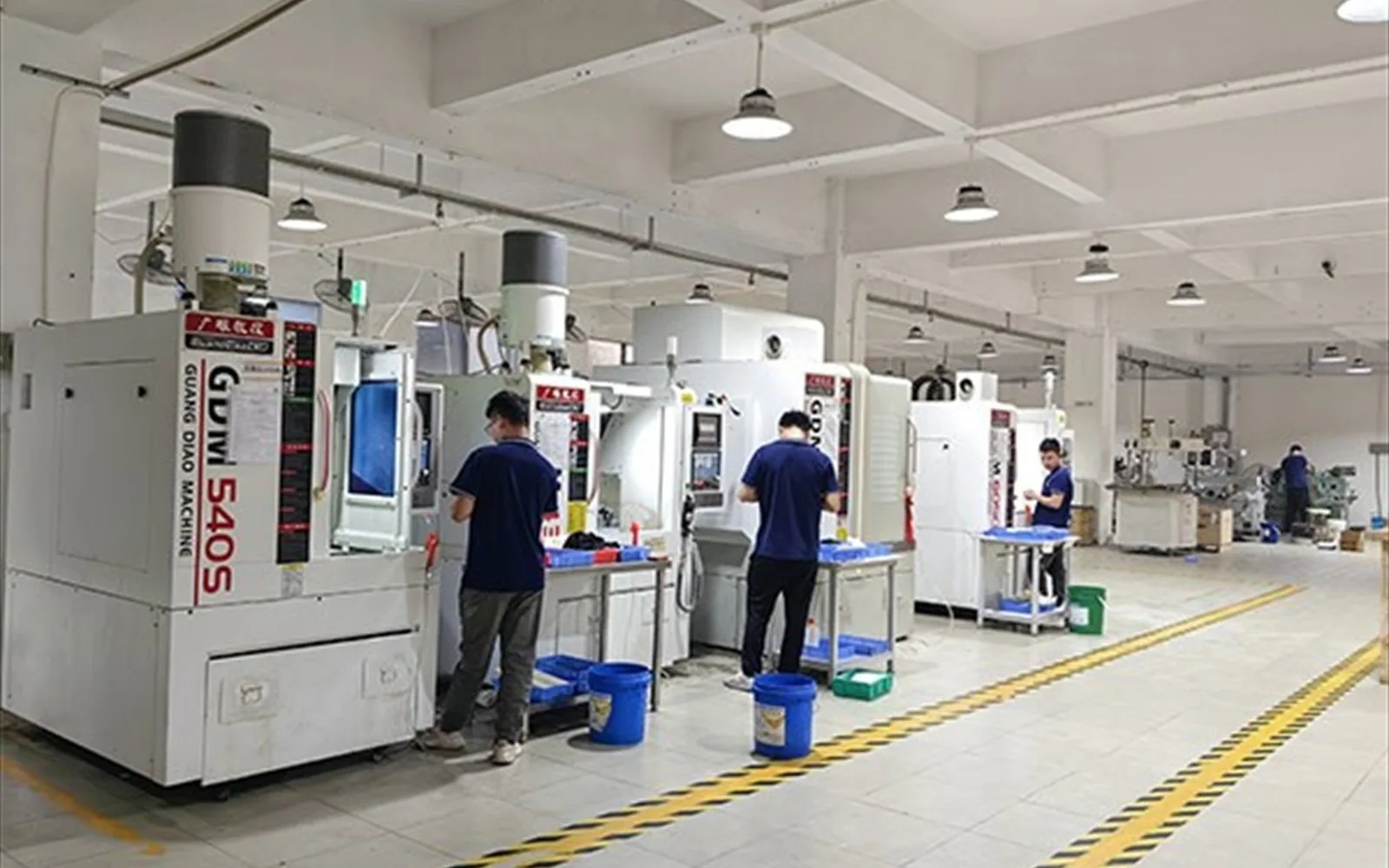 Three operators working at vertical CNC machining centres in a white machine hall, with blue drums beside the work tables and yellow-black marking on the floor.