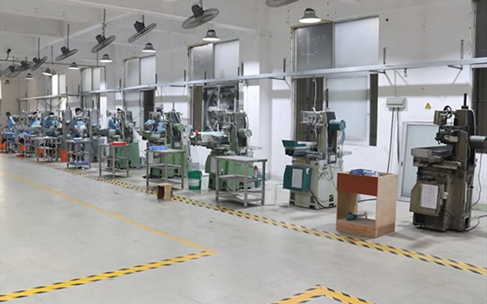 A long row of surface grinders along the Vintel@Ceramics shop floor, each at its own bench under pendant lights and ceiling fans, with yellow hazard markings on the floor.