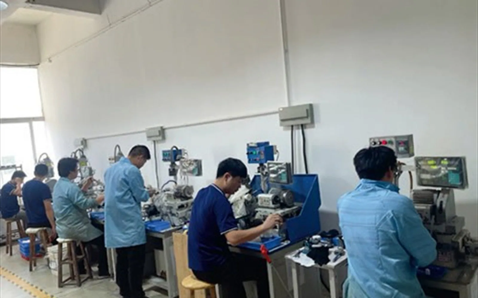 Technicians seated along a white wall working at lapping and polishing stations, each station fitted with a control box and a spindle head.