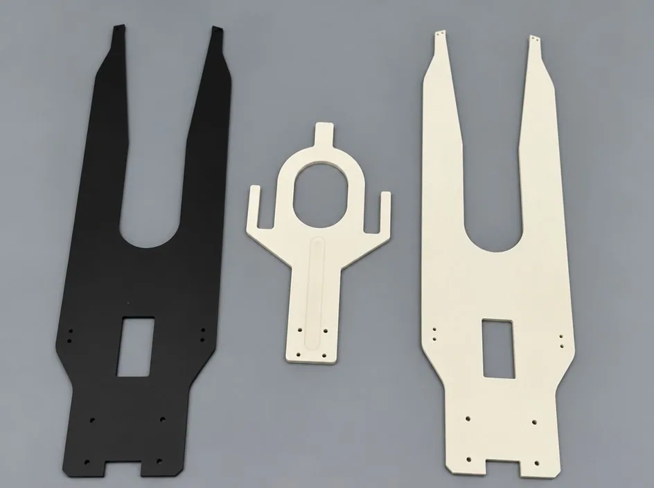 Three components on a grey studio ground: two large white alumina fork plates and a smaller machined part, beside a dark component of similar outline.