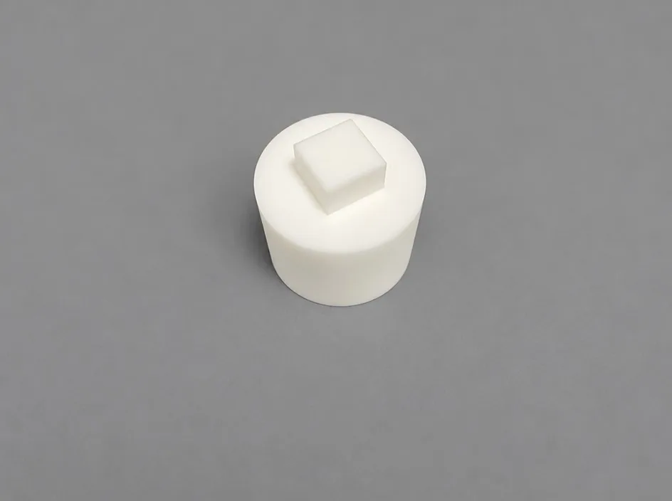 A cylindrical white body on a grey studio ground with a raised square boss on its top face.