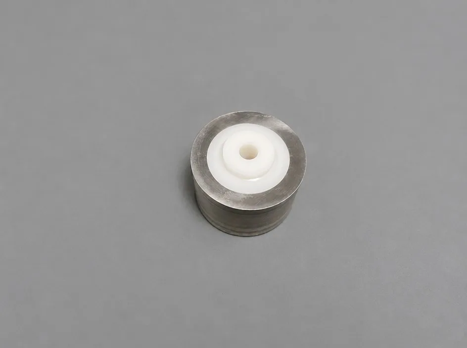 A shallow metal cup on a grey studio ground with a white ceramic insert centred in it, the insert carrying a small through bore and a raised ring.