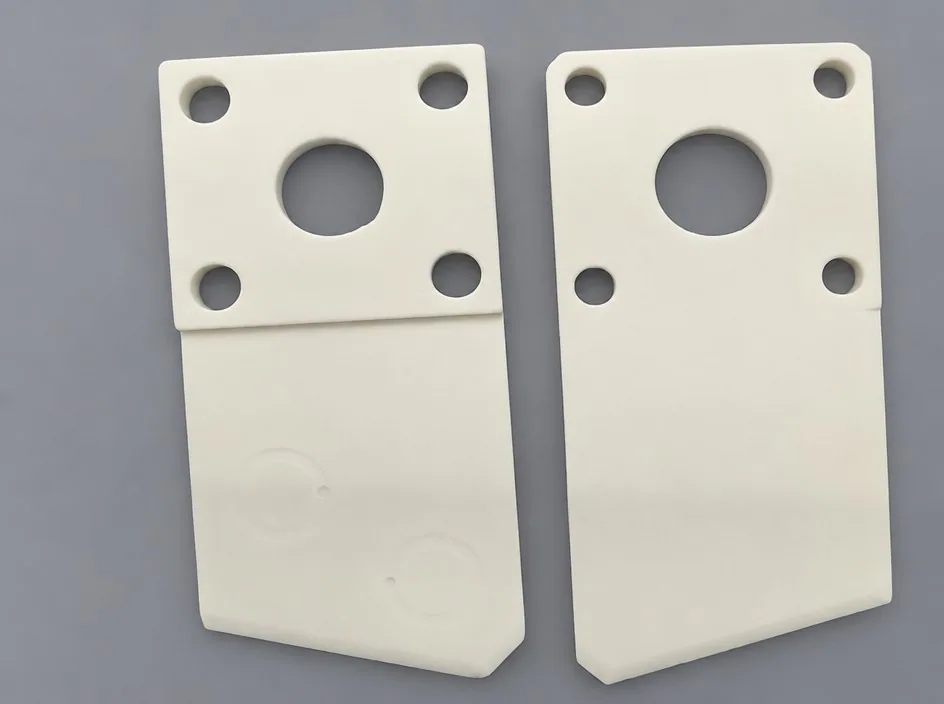 Two white plaques on a grey studio ground, each drilled with four corner bores and a larger central bore, and carrying a faint circular mark.
