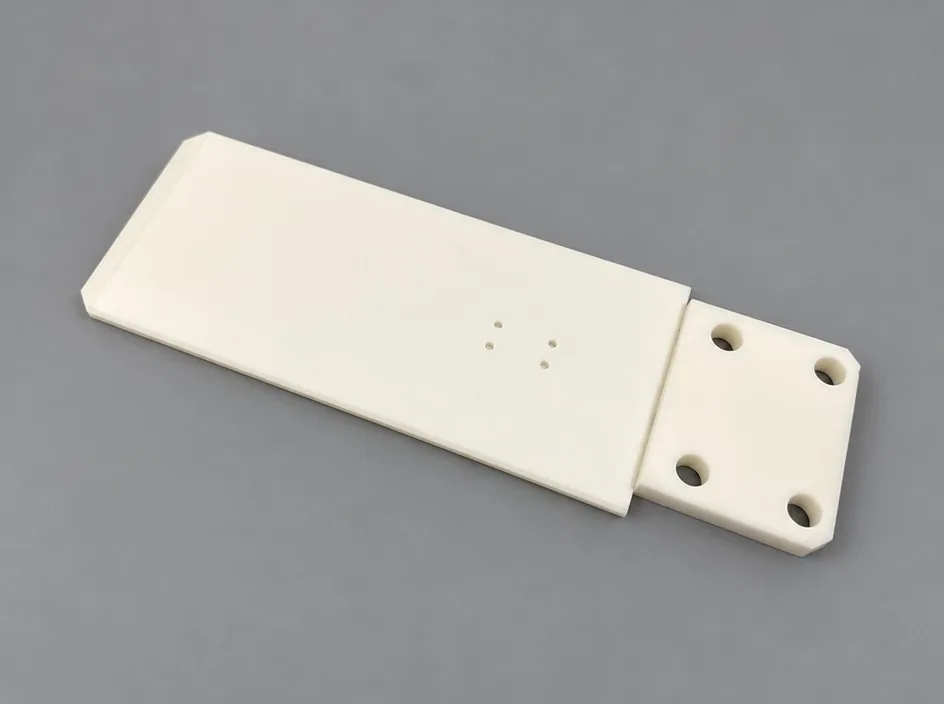 A white plate on a grey studio ground with a stepped and chamfered end carrying four bores, and a shallow circular mark on the body.